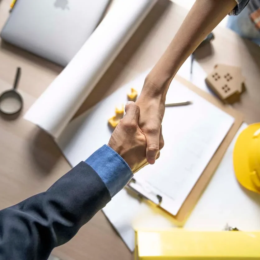 Two people shaking hands over a table with construction materials.